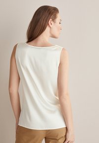 Beżowa bluzka bez rękawów o gładkiej fakturze i okrągłym dekolcie. Widok z tyłu ukazuje delikatne drapowanie i dopasowany krój, w połączeniu z beżowymi spodniami.