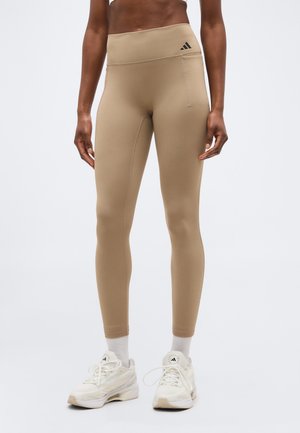 Μπεζ κολάν με ψηλή μέση και μικρή πλαϊνή τσέπη, φορεμένα με λευκές κάλτσες και λευκά παπούτσια Adidas σε όρθιο άτομο.