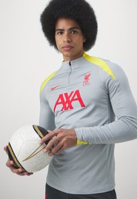 Harma pitkähihaine urheiluopas, jossa on keltaisia yksityiskohtia, korkea kaulus ja punainen Liverpool FC -logo, yhdistettynä teksturoituun valkoiseen jalkapalloon.