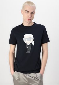 Fekete póló, amelyen egy figura látható, fehér hajjal és napszemüveggel, egy egyszerű háttér előtt. Az anyag simának tűnik.