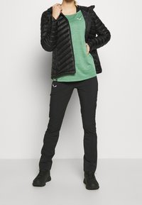 Černá prošívaná bunda přes zelený sportovní top, v kombinaci s černými kalhotami a tmavými botami. Má zipové zapínání a úzký střih.