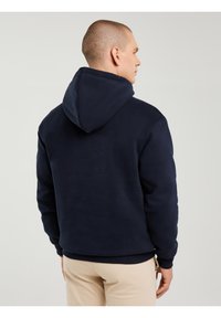 Vīrietis redzams no muguras, valkājot tumši zilu kapuci un bēšas bikses, uz vienkrāsaina gaiši pelēka fona.