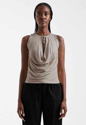 Jeune femme aux cheveux tressés portant un haut beige sans manches drapé noué au niveau du col et un pantalon noir à cordon de serrage.