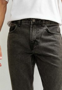 Primo piano di una persona che indossa jeans di denim neri e una maglietta bianca, con una mano sulla vita, su uno sfondo chiaro.