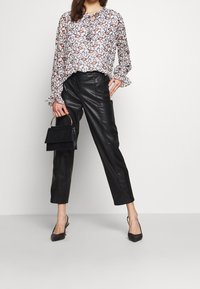 Doorzichtig wit blouse met een bruin en zwart bloemmotief, met gerimpelde manchetten; gecombineerd met hoge zwarte leren broek en zwarte hakken.