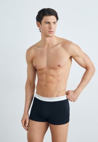 Schwarze Baumwoll-Boxershorts mit einem weißen elastischen Bündchen, das das "GANT"-Logo trägt. Das Modell hat einen muskulösen Körperbau und steht vor einem schlichten Hintergrund.