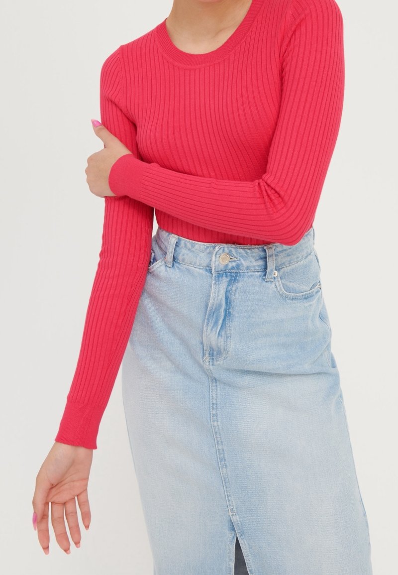 Femme portant un haut rouge vif à manches longues côtelé avec une jupe en denim bleu clair taille haute, le bras plié et la main touchant le bras opposé.