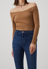 Hnědý žebrovaný top s dlouhým rukávem a designem bez ramínek, spárovaný s modrými džínami s vysokým pasem, které mají zapínání na knoflíky a jemné efekty vyblednutí.