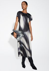 Frau trägt ein schwarz-weißes, gradient Midi-Kleid, hält eine große silberne Clutch und trägt schwarze Stiefeletten mit Absatz sowie Creolen.