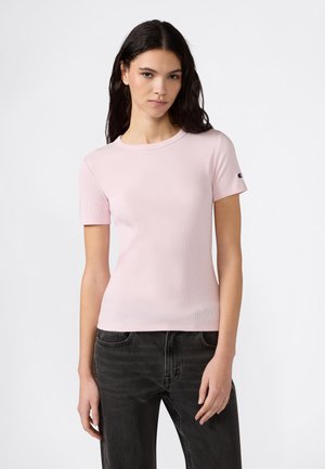 Mujer joven con cabello oscuro, vistiendo una camiseta de manga corta rosa claro con textura acanalada y jeans negros, posando frente a un fondo blanco liso.