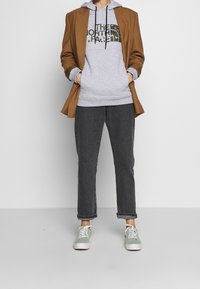 Sudadera gris con logo, blazer marrón, jeans oscuros y zapatillas verdes. Presenta cordones en la sudadera y jeans con puños. Material texturizado.