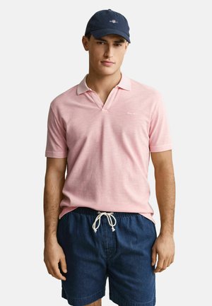 Homme portant un polo rose à manches courtes, un short bleu marine à cordon de serrage et une casquette bleu marine, debout devant un fond uni.