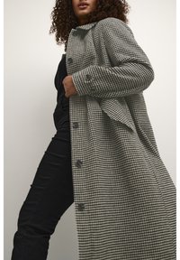 Lång kappa i houndstooth-mönster i grått och svart, med stora knappar, krage och avslappnad passform. Bärs över en svart outfit.