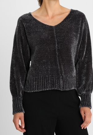 Femme portant un pull en velours côtelé gris foncé à manches longues et col en V avec un pantalon noir, vue du cou jusqu'aux hanches.