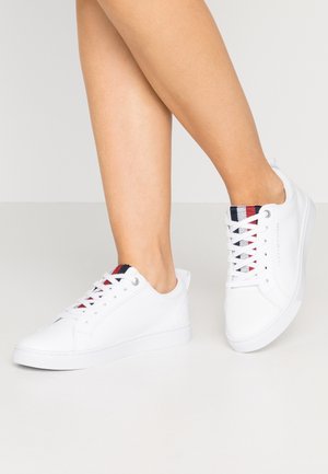 Beneden benen met witte lage sneakers met rode, marineblauwe en witte gestreepte details op de tong, staand op een effen lichte vloer.