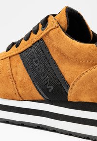 Zapatillas de ante en color mostaza con acentos de textil negro. Cuentan con cordones negros y una suela de rayas blanca y negra.