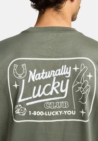 Camiseta verde oliva con un gráfico blanco en la parte posterior. El diseño incluye un herradura, un gesto de mano y el texto: "Club de la Suerte Natural".