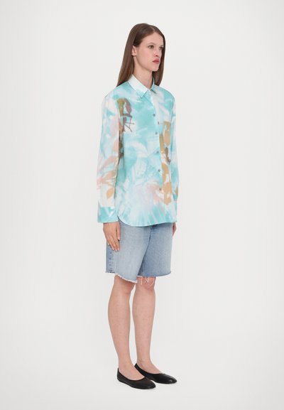 Lichtgewicht, langemouwen overhemden met een blauw en beige abstract patroon, gecombineerd met spijkerbroeken en zwarte flats.
