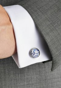 Bouton de manchette au design circulaire avec un motif floral bleu sur un tissu doux, posé sur une manche de chemise blanche avec un costume gris texturé.