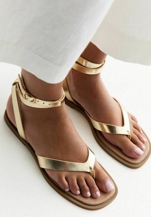 Piedi con sandali alla caviglia in metallo dorato con suole beige, abbinati a pantaloni bianchi a gamba larga, su sfondo bianco.