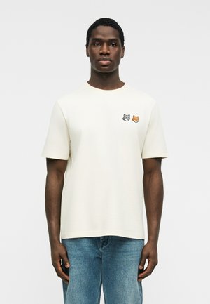 Άνδρας που φοράει άσπρο κοντομάνικο μπλουζάκι με γκρι και πορτοκαλί μπαλώματα κεφαλής αλεπούς στο αριστερό στήθος, συνδυασμένο με μπλε τζιν.