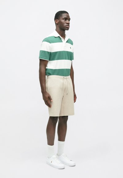 Πράσινο και λευκό ριγέ πουκάμισο πόλο, μπεζ σορτς με κορδόνι, λευκά αθλητικά παπούτσια και ανοιχτόχρωμες κάλτσες. Το μοντέλο στέκεται μπροστά σε ένα απλό φόντο.