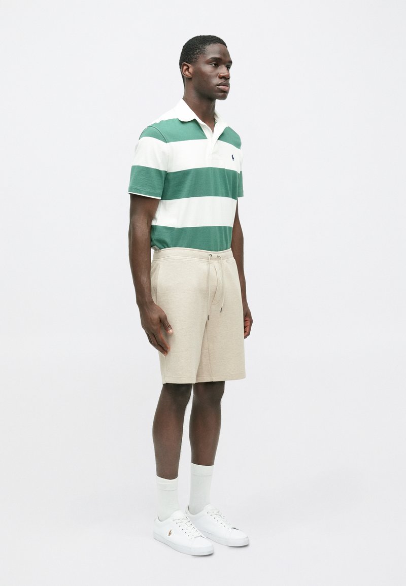Groen en wit gestreepte polo, beige shorts met koord, witte sneakers en lichte sokken. Model staat rechtop tegen een effen achtergrond.