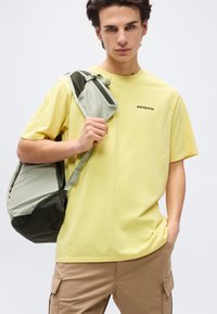 Νεαρός άνδρας που φοράει κίτρινη μπλούζα Patagonia και μπεζ παντελόνι cargo, κρατώντας λουρί γκρι και μαύρου σακιδίου στον ώμο του.