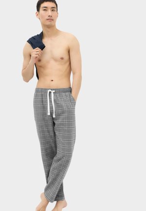 Junger Mann steht barfuß, trägt graue karierte Pyjamahose mit weißem Kordelzug und hält ein dunkles Hemd über der Schulter vor einem einfarbigen Hintergrund.