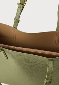 Žalios tekstūruotos odinės rankinės su užtrauktuku atsidarančiu rudu vidumi. Turi ilgus rankenas ir matomus siūlių siuvimo akcentus.