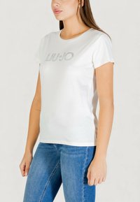 Biała bawełniana koszulka z okrągłym dekoltem, krótkimi rękawami i srebrnym logo "LIU JO" z kryształkami na przodzie. Dobranie do niebieskich dżinsów.