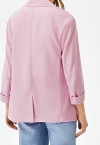 Pink blazer with a relaxed fit, long sleeves, and rolled cuffs. Smooth texture with a vented back and a classic lapel collar.