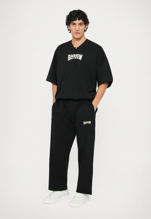 T-shirt nero a maniche corte e pantaloni abbinati, entrambi con il logo bianco "BARROW". Il modello indossa sneakers bianche. Vestibilità semplice e rilassata.