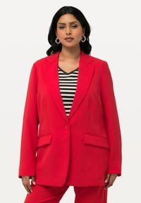 Blazer rosso con rever, un bottone e due tasche davanti; abbinato a un top a righe bianco e nero. Tessuto di texture liscia.