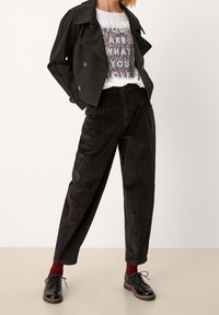 Žena v černém zkráceném dvojitě zapínacím saku, tištěném bílém tričku, černých kalhotách s vysokým pasem, červených ponožkách a černých kožených botách.