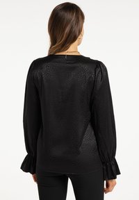 Sort bluse med tekstureret mønster, lange ærmer med let rynkede manchetter og en let glans. Lukning bagpå med en lille knapdetalje.