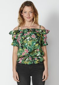 Top floreale nero con spalline abbondonate, caratterizzato da foglie verdi e fiori rosa, bordi arricciati e fascia elastica in vita. Abbinato a jeans scuri.