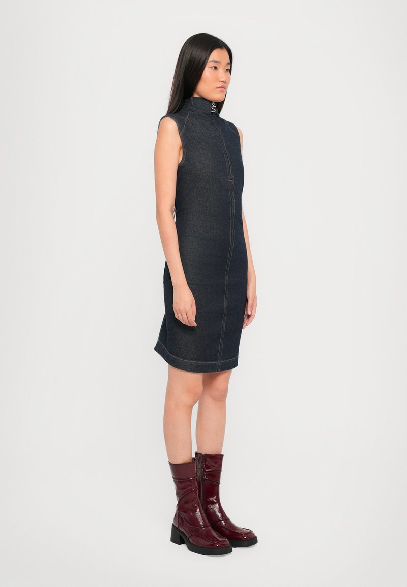 Robe en denim sans manches avec un col haut, fermeture éclair frontale et surpiqûres contrastées, associée à des bottines brillantes bordeaux à talon épais.