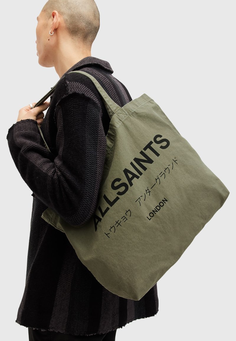 Borsa tote in tela verde oliva con testo nero "ALLSAINTS LONDON" e un testo giapponese, portata a spalla da una persona in un maglione nero.