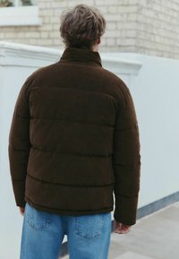 Brun kordfløjl pufferjakke med høj krave og quiltet design. Bæres med lyseblå jeans. Glat tekstur, afslappet pasform og ingen synligt hardware.