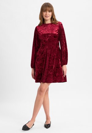 Rotes Samtkleid mit langen, gepufften Ärmeln und rundem Halsausschnitt. Die strukturierte Stoffqualität verleiht einriches Aussehen und fällt über das Knie. Es werden schwarze Ballerinas getragen.