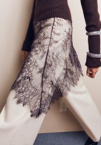 Osoba oblečená v hnedom žebrovanom svetri, krémových širokých nohaviciach a priehľadnej čiernej čipkovanej sukni s kvetinovými vzormi.