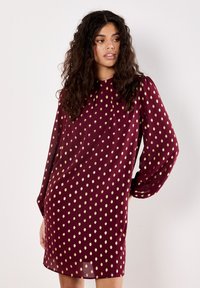 Donna con lunghi capelli ricci che indossa un abito bordeaux con lucidi pois dorati, in piedi davanti a uno sfondo bianco uniforme.