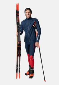 Längdskidåkare i en marinblå jacka, röd och blå tävlingsdräkt samt svarta stövlar. Han håller skidor och stavar med röda detaljer.