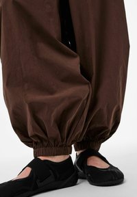 Osoba ubrana w luźne brązowe spodnie z elastycznymi mankietami i czarne, zakryte buty na rzepy o szerokich paskach, stojąca na białej powierzchni.