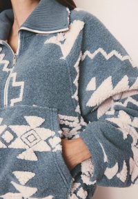 Fleece-paita, jossa on vetoketjukaulus, sinisellä pohjalla ja valkoisilla geometrisilla kuvioilla. Huomattava pörröinen rakenne ja etutasku.