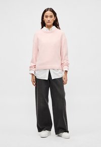 Ροζ ανοιχτόχρωμο πλεκτό πουλόβερ με στρογγυλή λαιμόκοψη, συνδυασμένο με λευκό πουκάμισο με γιακά και μαύρο τζιν παντελόνι με φαρδιά μπατζάκια. Το σύνολο ολοκληρώνουν λευκά αθλητικά παπούτσια.