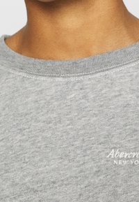 Harmaa collegepaita teksturoidulla kankaalla, pyöreällä pääntiellä ja vasemmassa rinnassa valkoisella kirjailulla "Abercrombie New York" -logo.