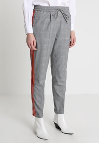 Šedé kostkované kalhoty s nastavitelným pasem, se červenými bočními pruhy a zúženým střihem. Nosené s bílými špičatými kotníkovými botami.