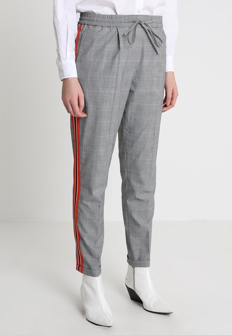 Šedé kostkované kalhoty s nastavitelným pasem, se červenými bočními pruhy a zúženým střihem. Nosené s bílými špičatými kotníkovými botami.